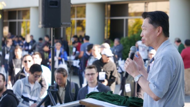 A speaker with a microphone addressing a large standing crowd outdoors.