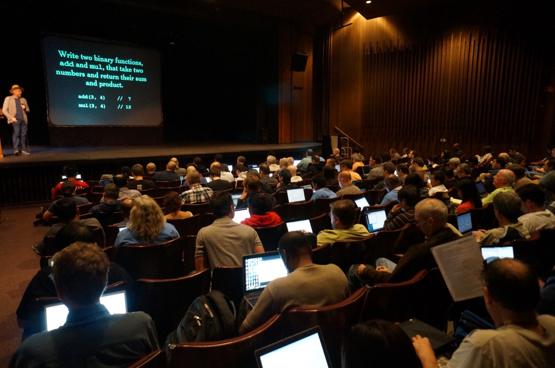 A packed auditorium of developers with laptops open, watching a speaker on stage beside a slide of code.