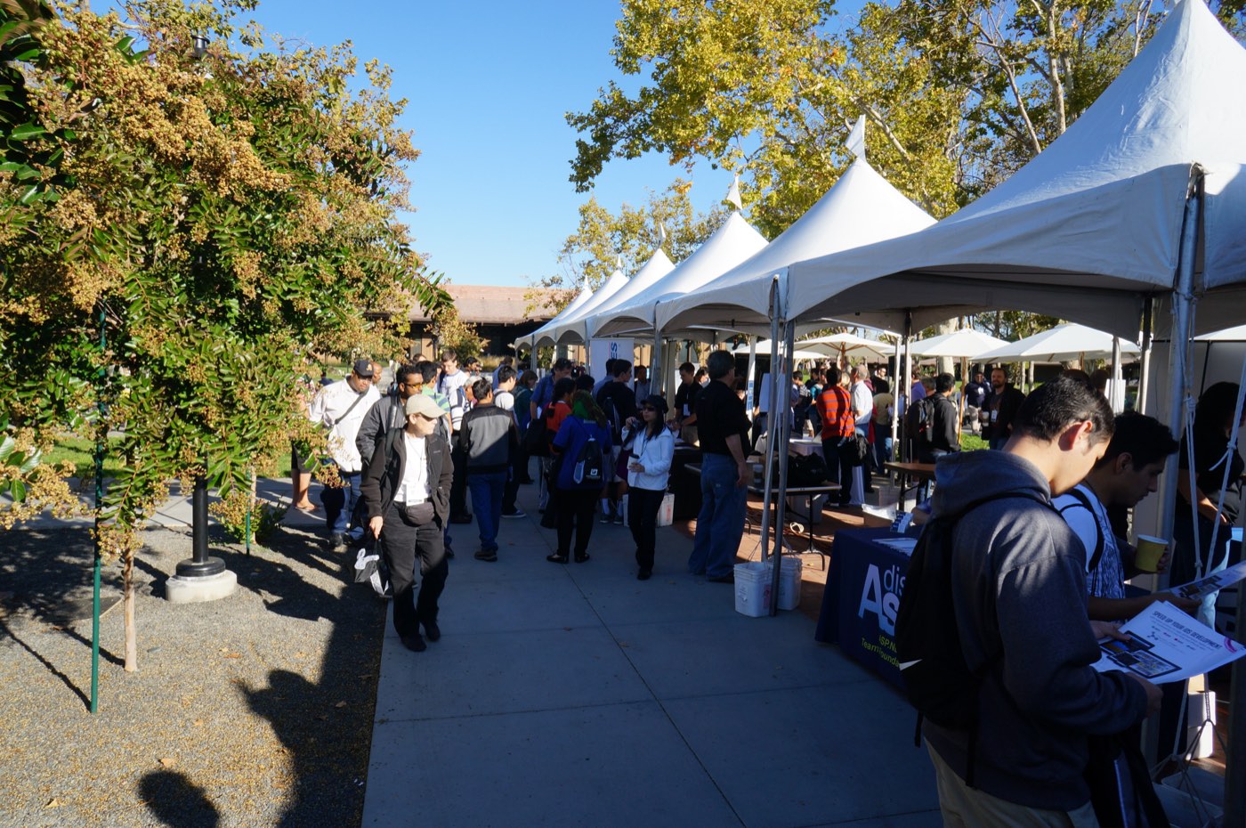 People walking past a row of white sponsor tents under trees on a sunny day.